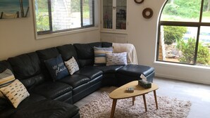 A living room with a black leather sectional sofa, a wooden coffee table, a fluffy rug, a glass cabinet, and a large window with a view of greenery. - Bayview water front holiday rental. (Lemon Tree Passage)