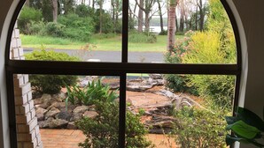 A room with a large arched window overlooking a landscaped garden with a pond, rocks, and various plants. - Bayview water front holiday rental. (Lemon Tree Passage)