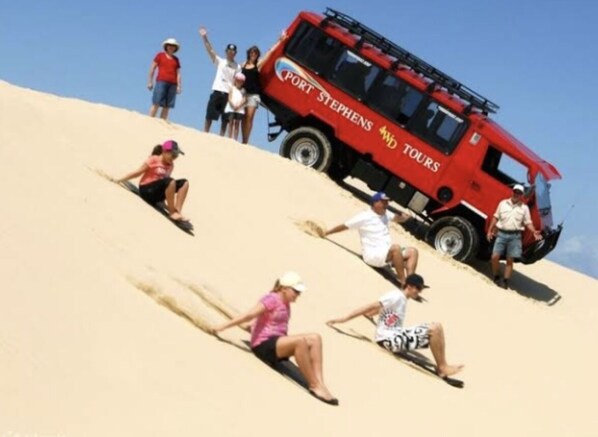 A group of people sliding down a sandy slope with a red vehicle labeled "Port Stephens Tours" in the background. - Bayview water front holiday rental. (Lemon Tree Passage)