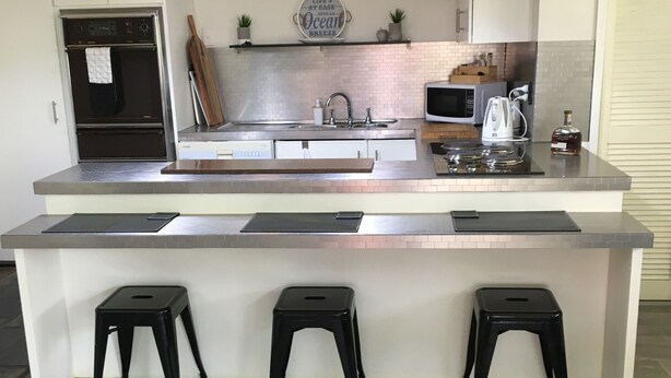 A modern kitchen with a central island, black bar stools, pendant lighting, and a stainless steel sink. - Bayview water front holiday rental. (Lemon Tree Passage)