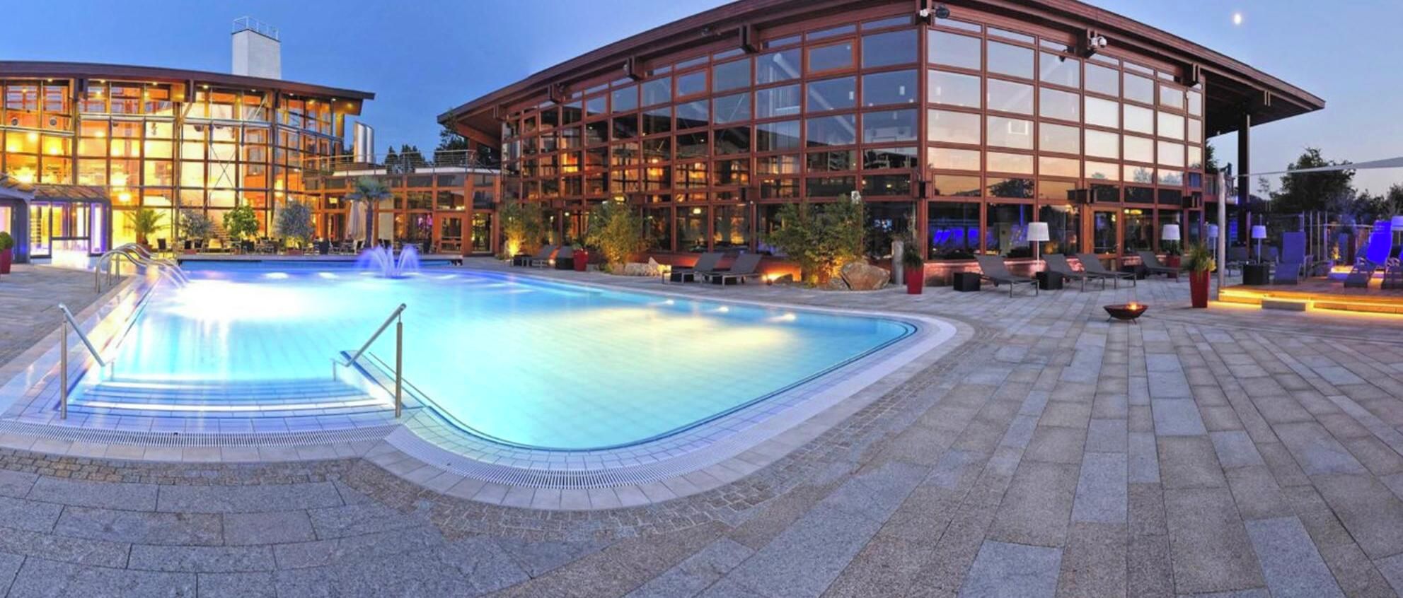 Ein Schwimmbecken mit einer Fontäne, umgeben von einer gepflasterten Fläche mit Liegestühlen und einem Gebäude mit großen Fenstern.
