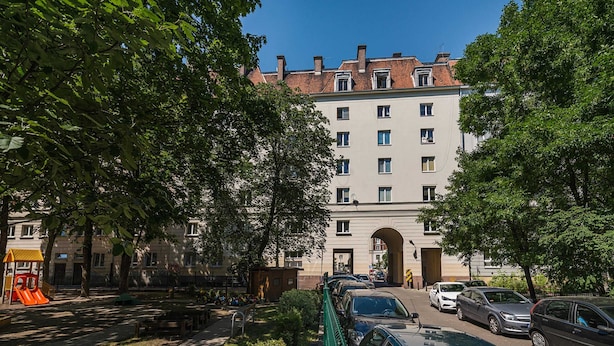 A multi-story building with a central archway entrance, surrounded by parked cars and a playground with a slide. - At the Fountain Apartment (Warsaw)