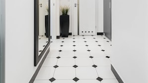 A hallway with white walls, a black and white tiled floor, a mirror on the left, a potted plant, and a door at the end. - At the Fountain Apartment (Warsaw)