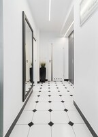 A hallway with white walls, a black and white tiled floor, a mirror on the left, a potted plant, and a door at the end.