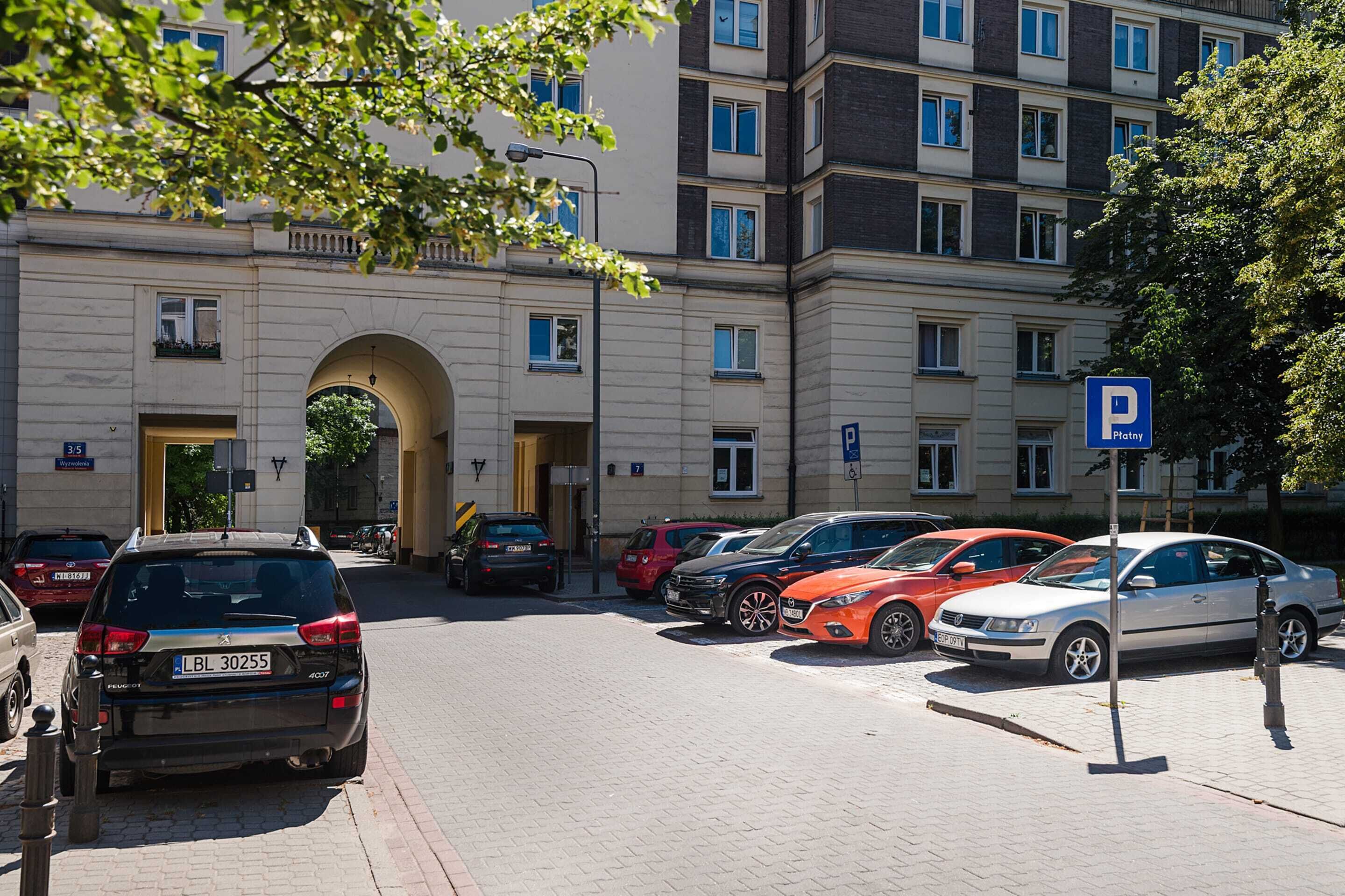 A parking area with multiple cars, a building with an archway entrance, and a blue parking sign.