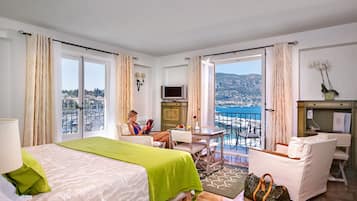 Ein Hotelzimmer mit Bett, Schreibtisch, Stuhl, einem Balkon mit Blick auf das Meer und die Stadt sowie einem Fernseher.