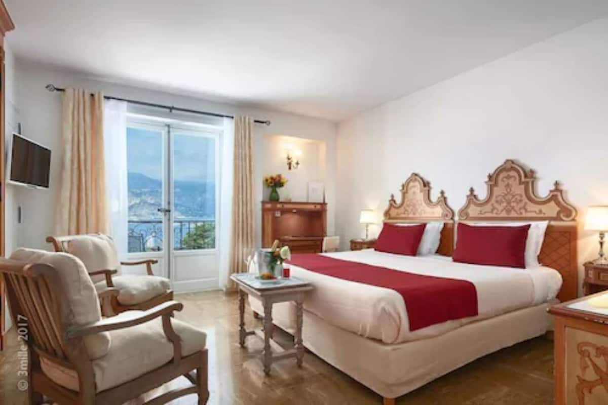 A hotel room with a large bed, a wooden chair, a small table, and a TV on the wall.