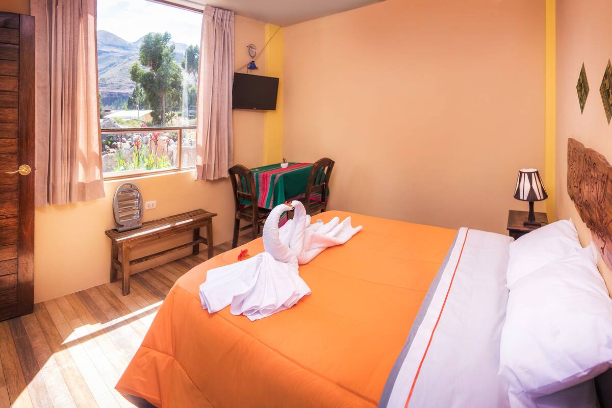 Una habitación de hotel con cama, un banco, una mesa con mantel colorido, una silla, una ventana con cortinas y vistas a las montañas.