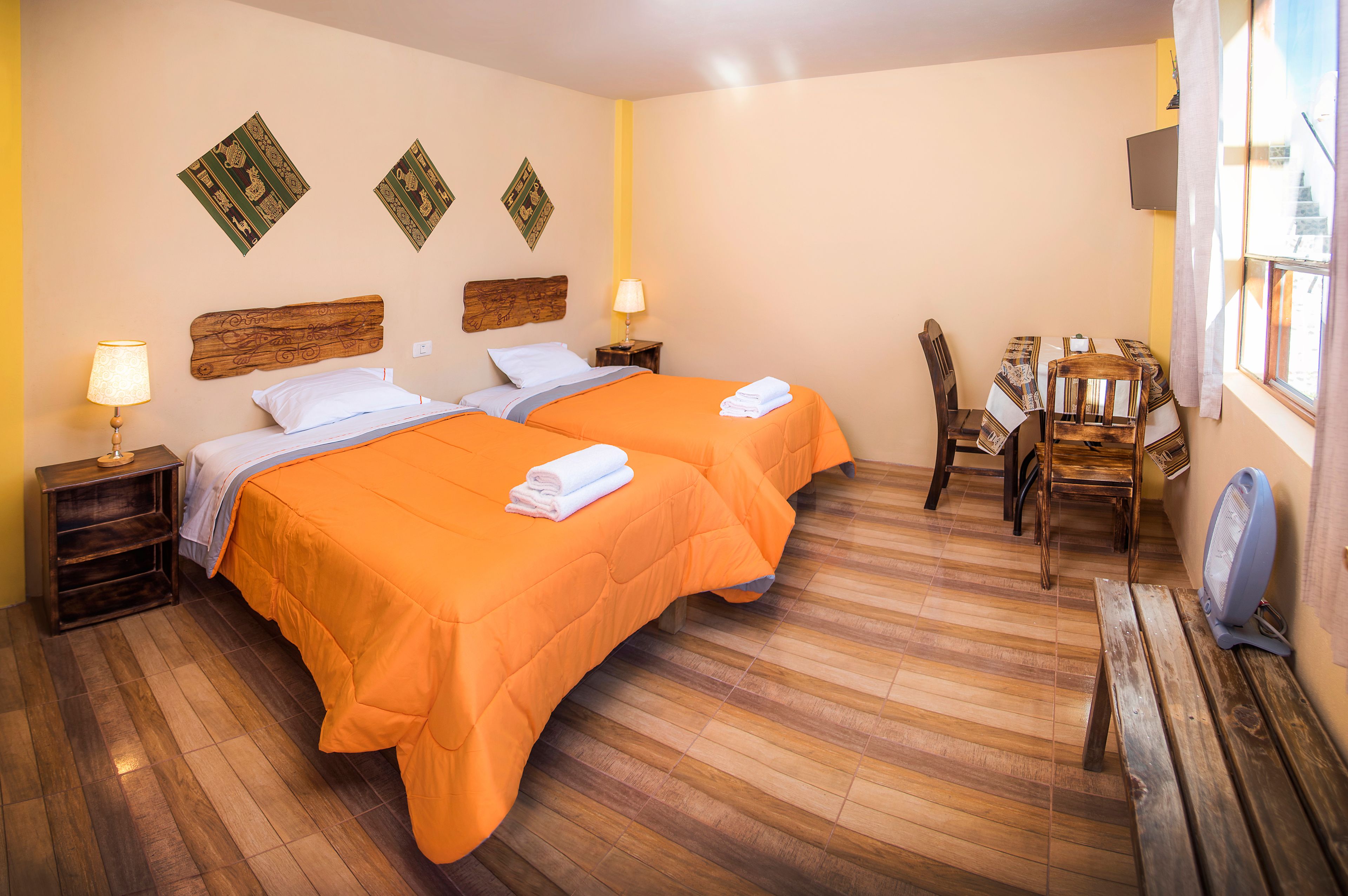 A hotel room with two beds, wooden headboards, a small table with chairs, a TV on the wall, and a wooden bench.