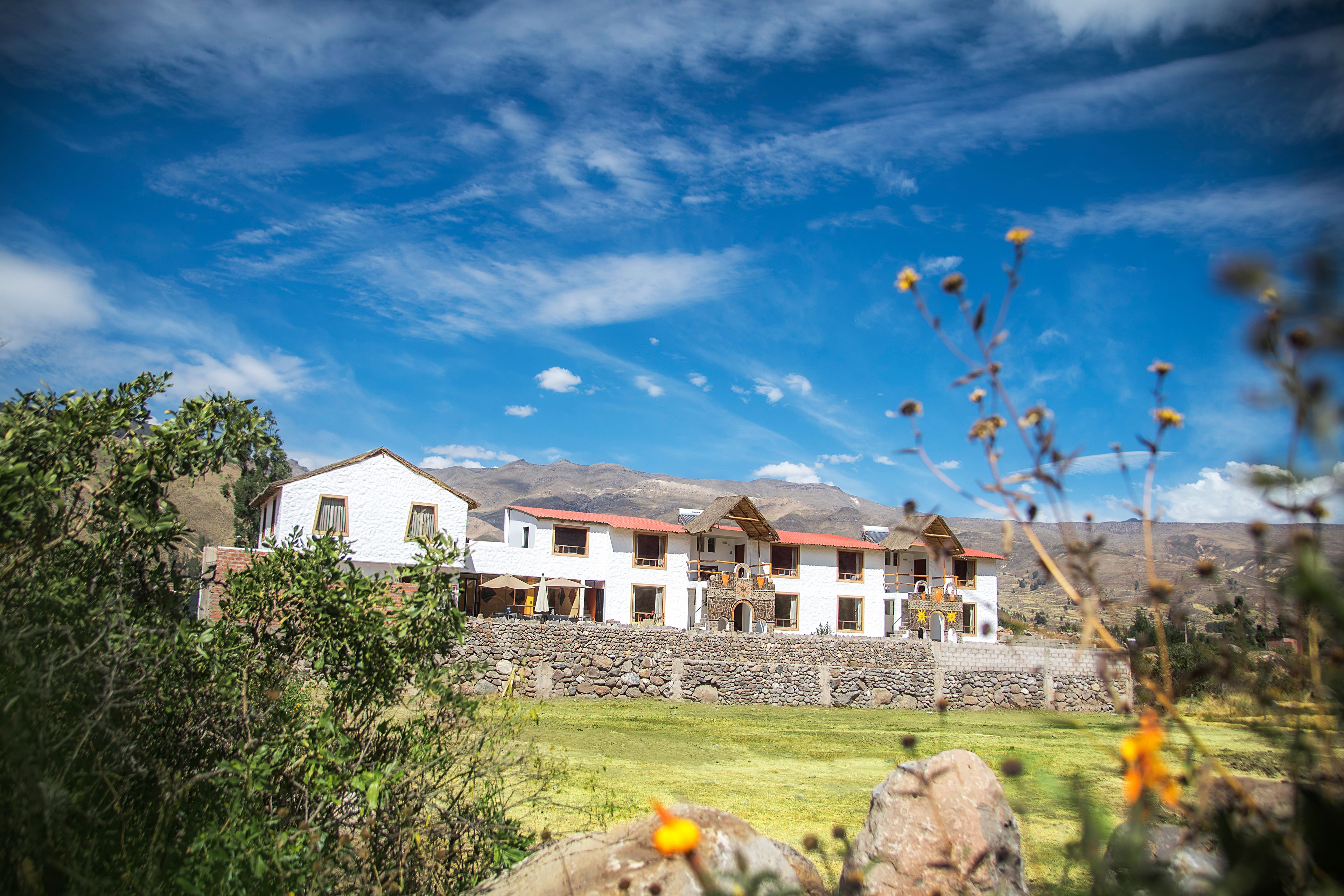 Un edificio blanco con numerosas ventanas y techo rojo, rodeado de vegetación y montañas al fondo.