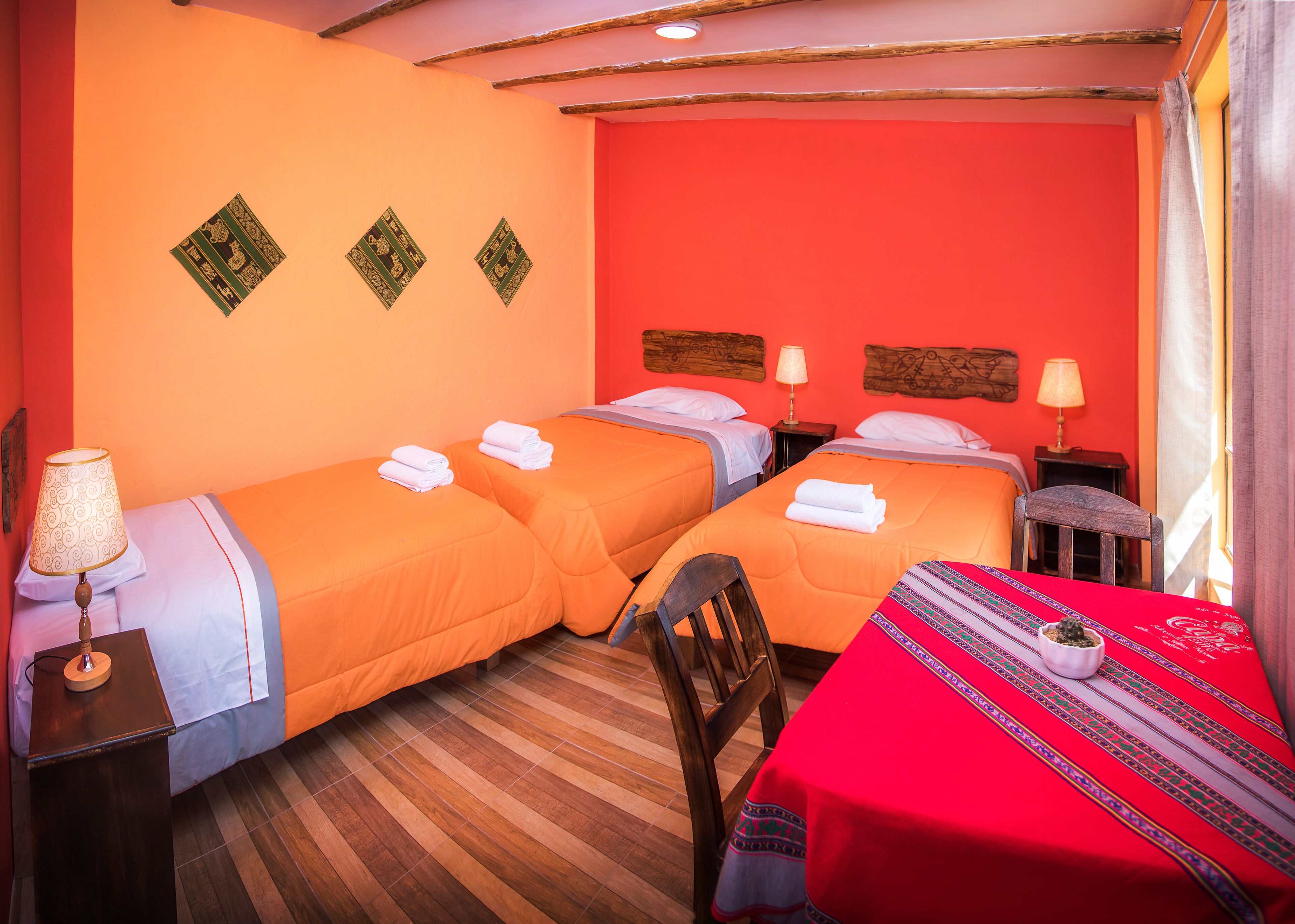 Una habitación de hotel con dos camas, una silla de madera, una mesa con mantel rojo y una lámpara en la mesita de noche.