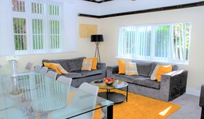 Un salon moderne avec deux canapés gris, une table basse en verre et une table à manger avec des chaises. De grandes fenêtres sont équipées de stores blancs, et on y trouve également une lampe sur pied et un tableau accroché au mur.