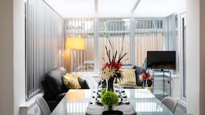 Ett modernt matområde med ett glasbord, svarta stolar, en soffa med kuddar, en tv och ett fönster med persienner.