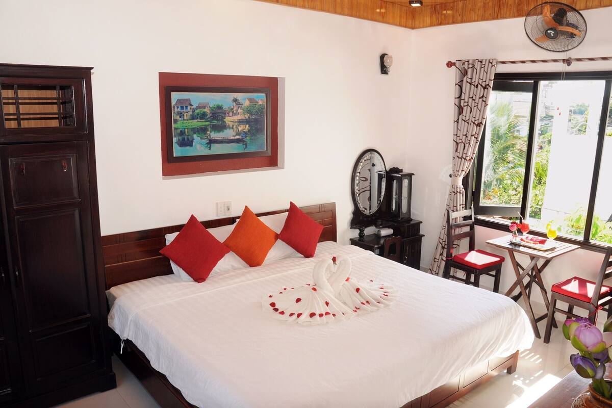 A hotel room with a bed, a painting, a desk with a chair, and a window with a view.