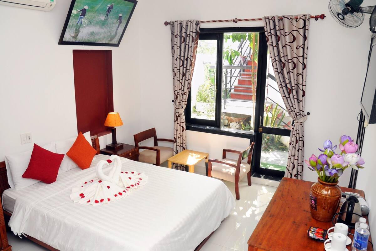 A hotel room with a bed, a desk, a chair, a table, a vase with flowers, and a window with curtains.