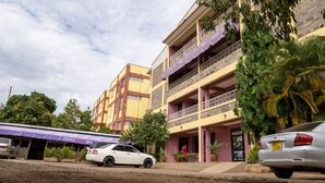 Un edificio de varios pisos con balcones, un coche blanco estacionado y un edificio con toldo morado.