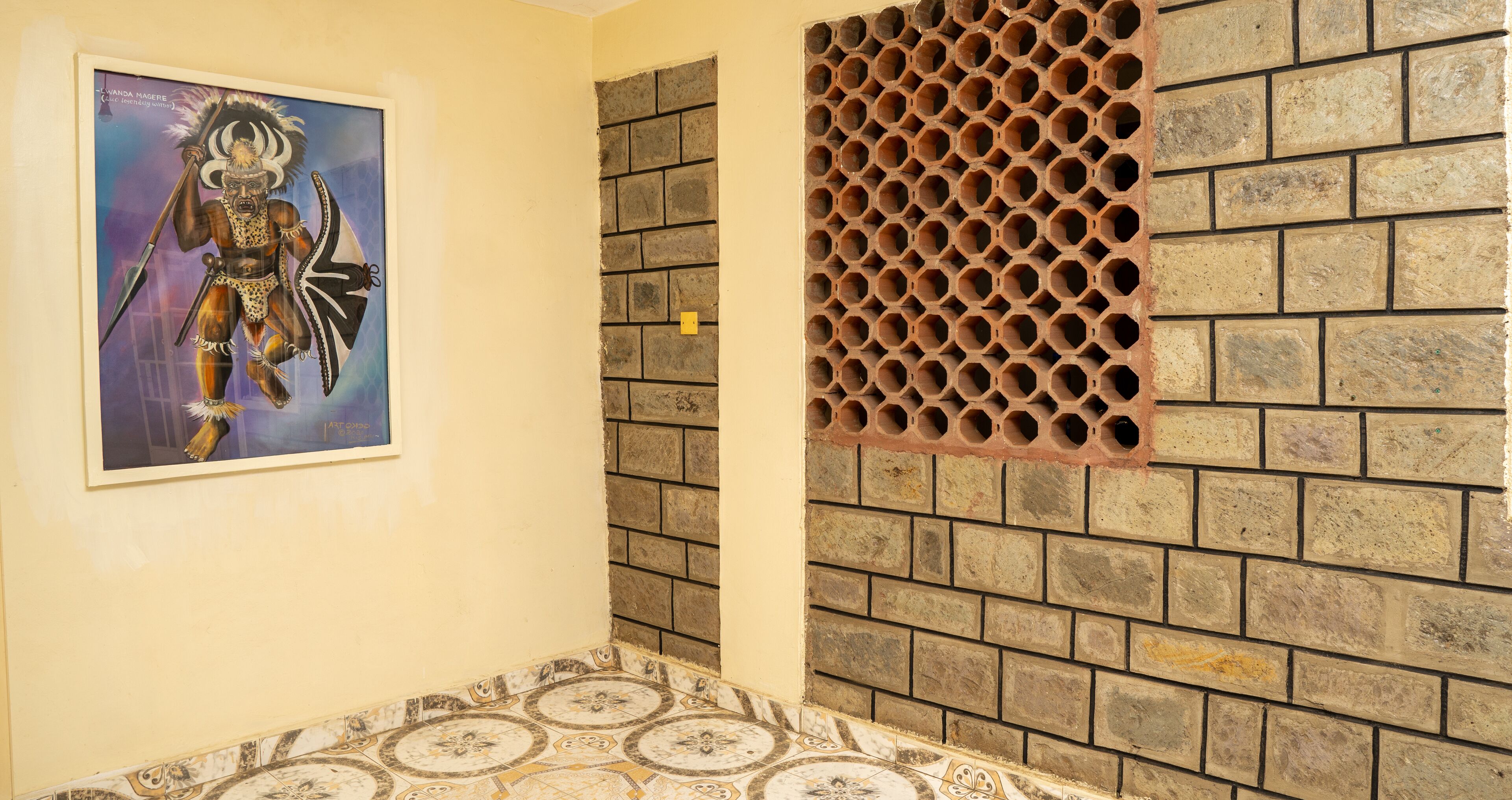 A room with a tiled floor, a brick wall with a decorative metal grille, and a framed artwork on the wall.