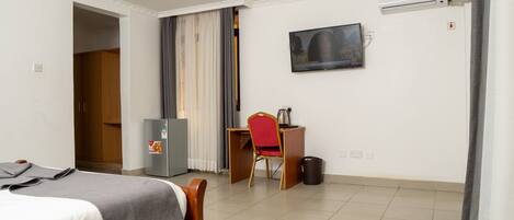 A hotel room with a bed, a desk with a chair, a refrigerator, a television, and an air conditioning unit.