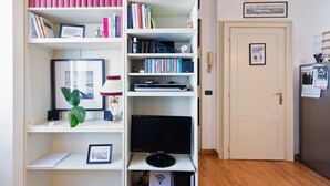 A room with a bookshelf containing books, a framed picture, and a small plant. There is a door with a picture on it and a wall-mounted shelf with various items. - Flatty Apartments Garigliano (Milan)