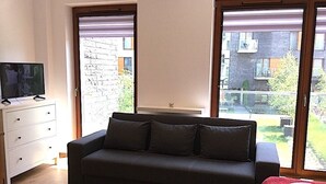 Huoneessa on tumma sohva, valkoisessa kaapissa oleva televisio ja suuri ikkuna, josta avautuu näkymä tiilitaloon ja vehreään maisemaan.