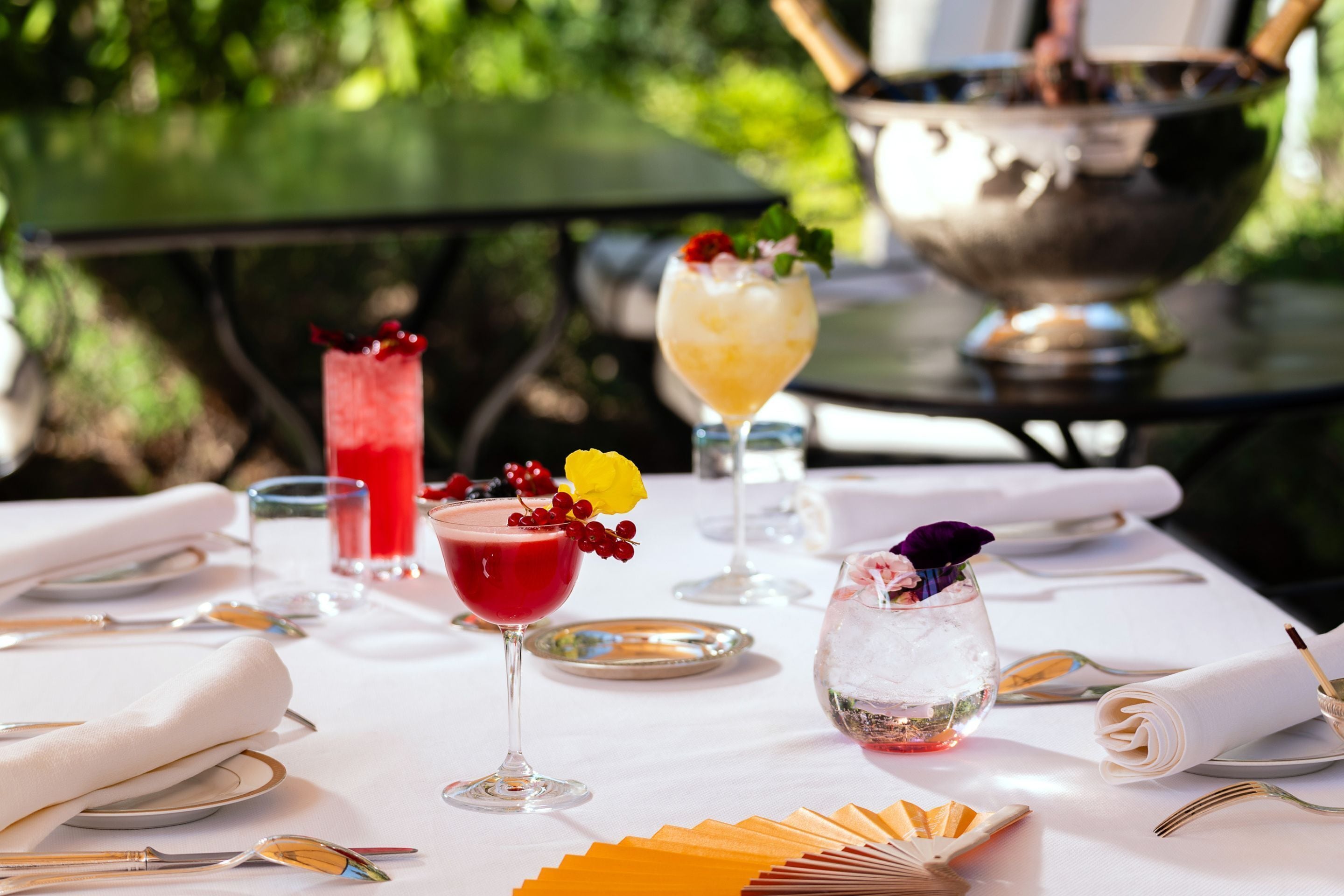 Une table dressée pour un repas, avec diverses boissons, un saladier en argent et un décor extérieur verdoyant.