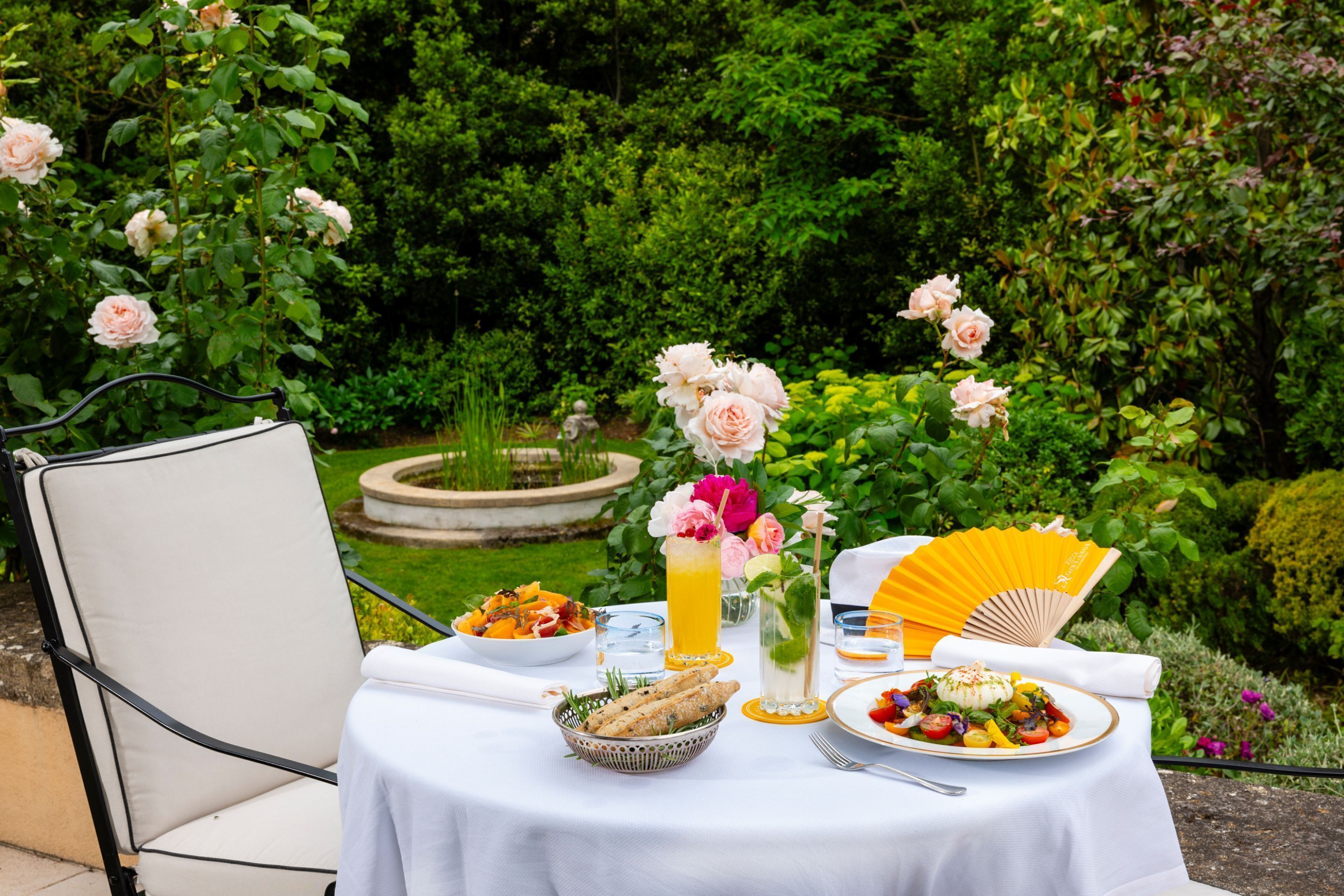Mesa de comedor al aire libre dispuesta con comida y bebidas, rodeada de follaje y flores.