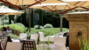 An outdoor seating area with tables and chairs under umbrellas, a small pond with lily pads, and a building in the background.