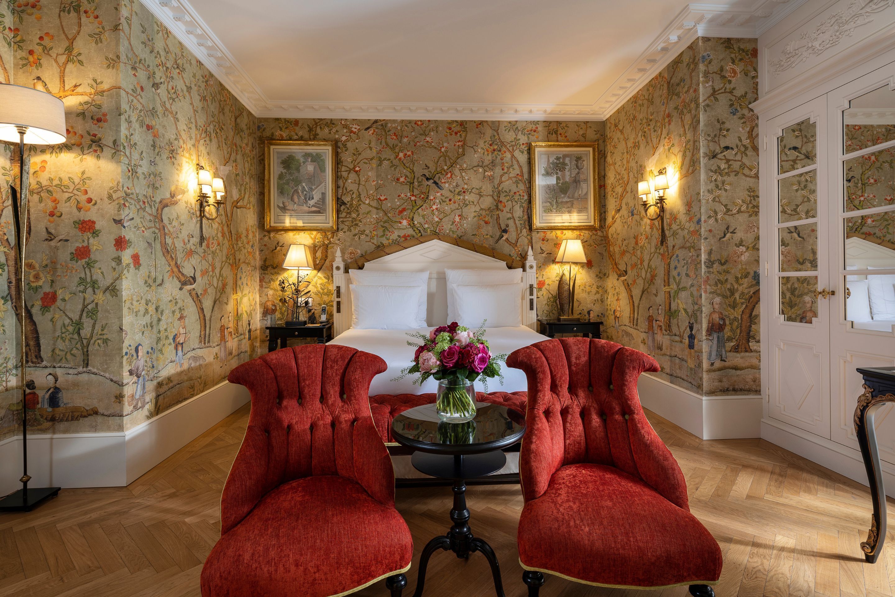 Una habitación con una cama, dos sillones rojos, una mesa redonda con un florero y papel tapiz floral.