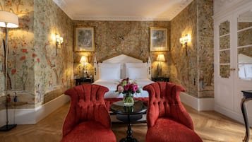 Una habitación con una cama, dos sillones rojos, una mesa redonda con un florero y papel tapiz floral.