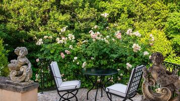 Un patio al aire libre con una mesa y dos sillas, rodeado de abundante follaje y flores rosadas.