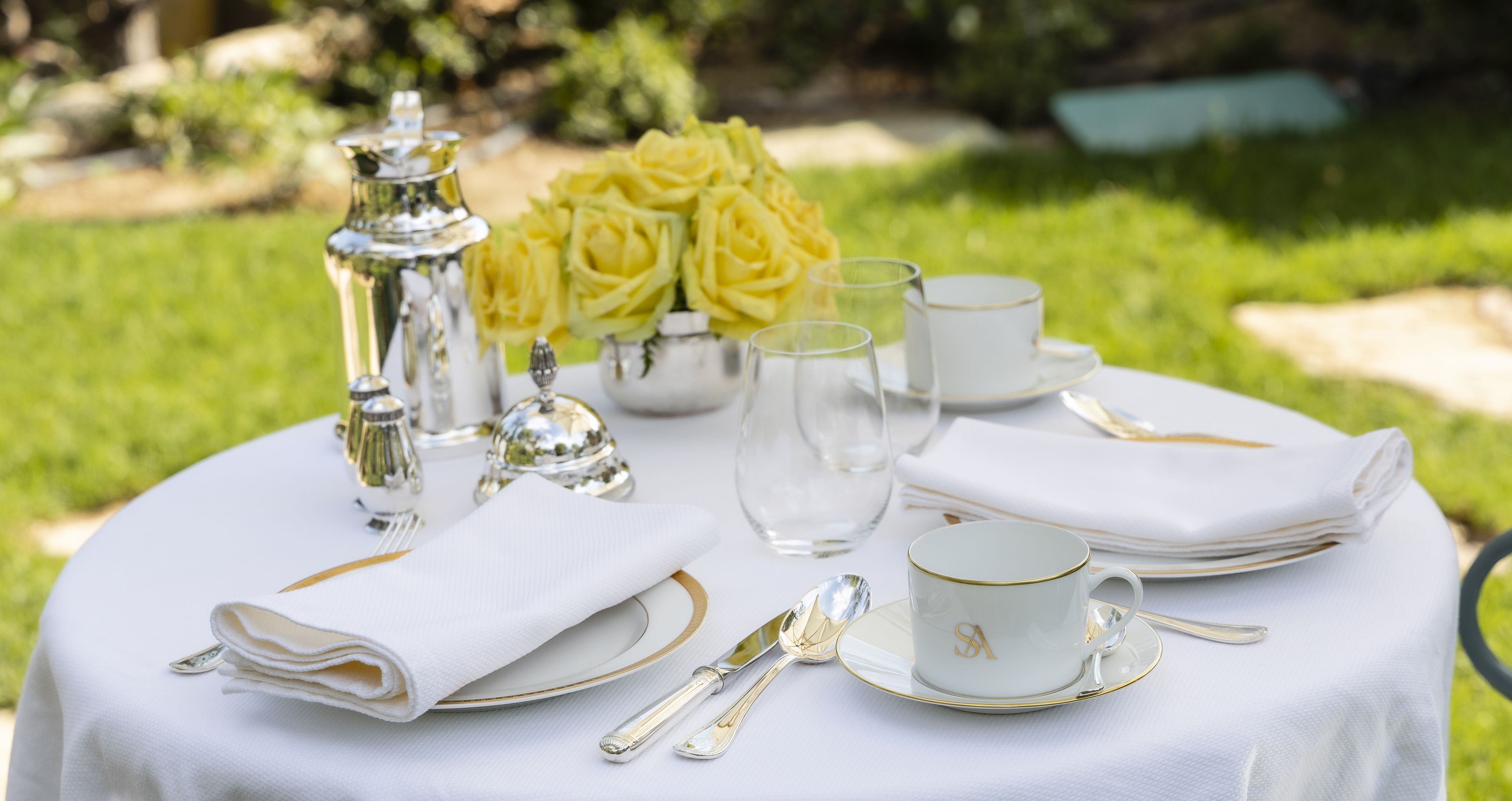 A round table set with white and gold tableware, including plates, cups, and cutlery, with a bouquet of yellow roses in the background.