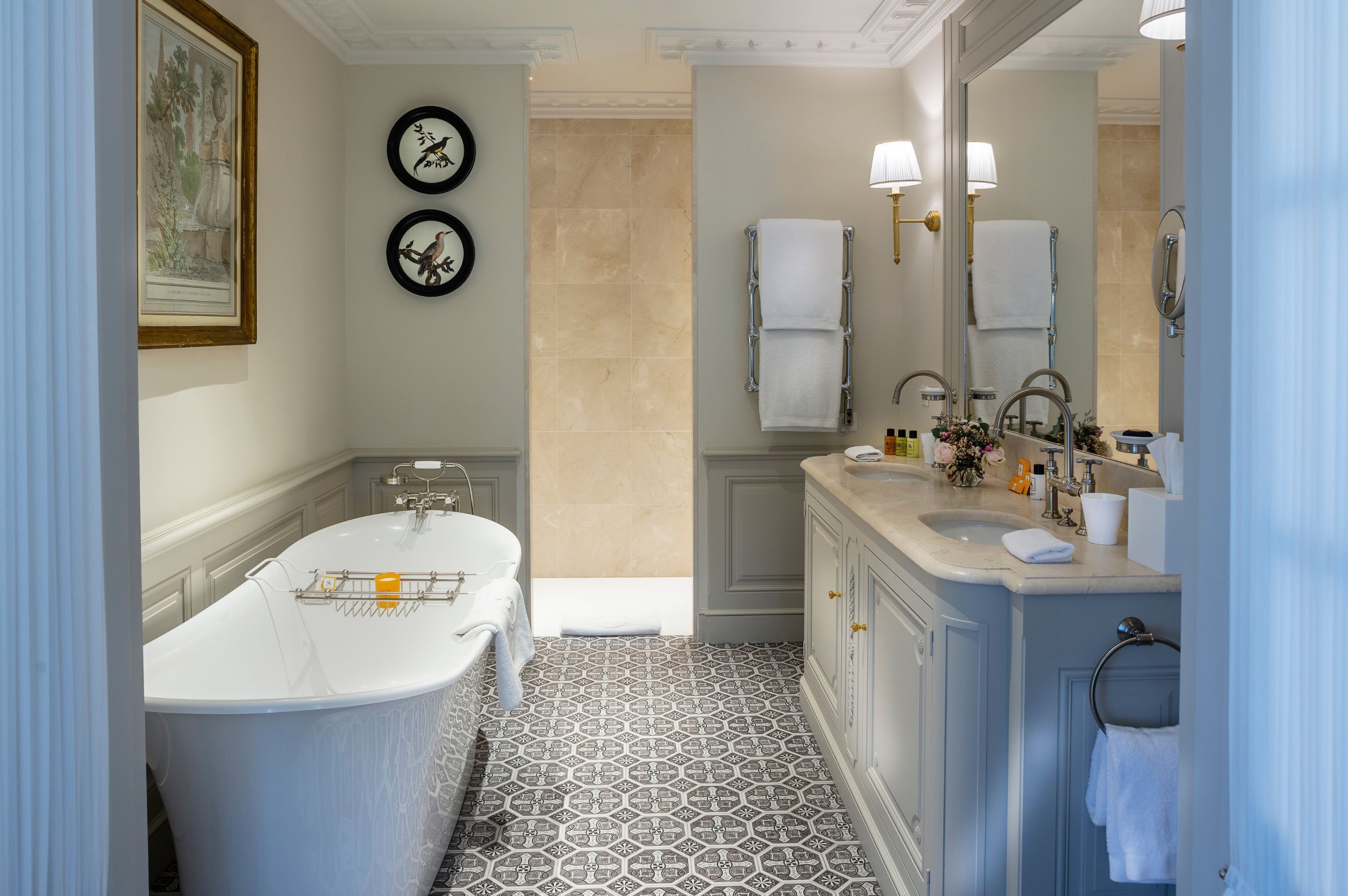 A bathroom with a freestanding bathtub, a vanity with a sink, and a large mirror. There are two wall-mounted clocks and a framed botanical print.