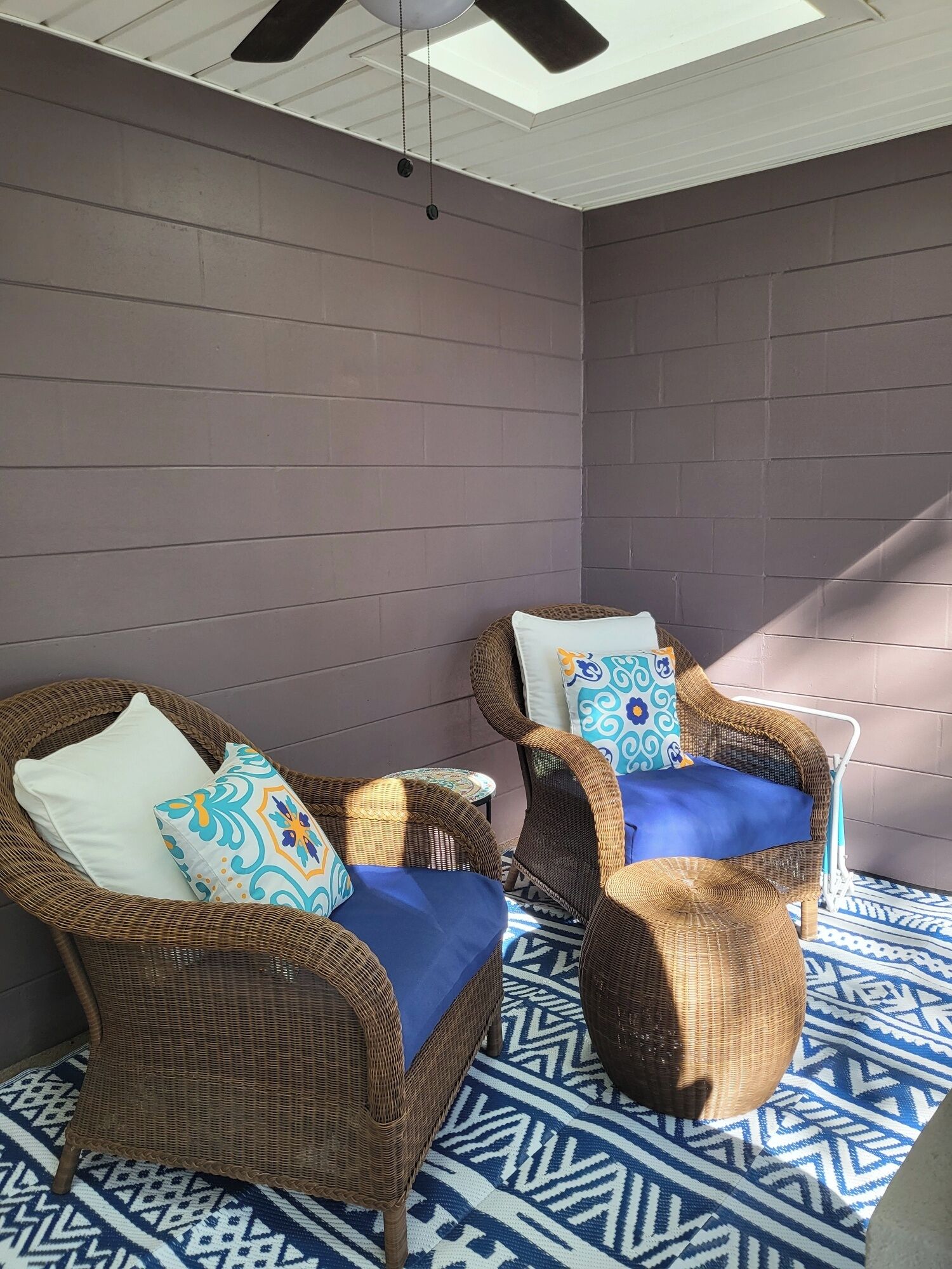 Deux fauteuils en osier avec des coussins bleus et des oreillers à motifs, un tapis à motifs et un ventilateur de plafond.