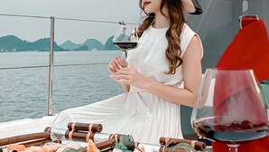 Eine Person auf einem Boot hält ein Weinglas, im Hintergrund ist Wasser und eine Berglandschaft zu sehen.
