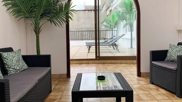 Une pièce avec une table à plateau en verre, deux canapés en osier ornés de coussins à motifs, une grande plante en pot et une vue sur un balcon extérieur avec une piscine et des chaises longues.