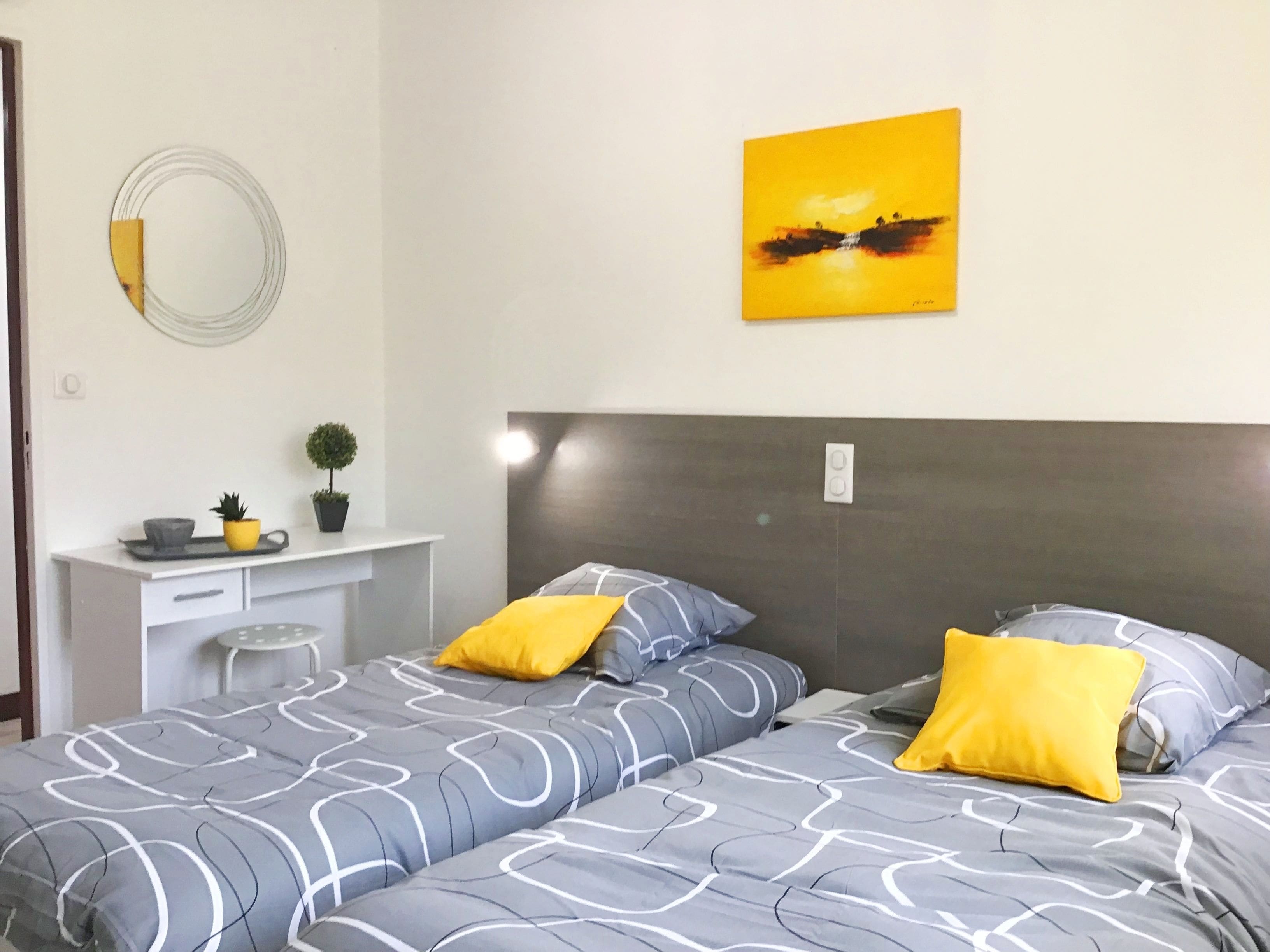 Une chambre moderne avec une couvre-lit grise à motifs, deux coussins jaunes, une tête de lit grise, un petit bureau avec une plante et un tableau jaune accroché au mur.