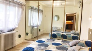 Une chambre avec un grand miroir, un lit avec une literie à motifs, un radiateur et une fenêtre ornée de rideaux légers.