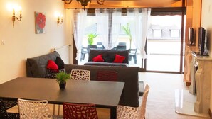 Un coin repas avec une table, des chaises et un lustre. On y trouve un canapé avec des coussins rouges et une plante posée sur la table.
