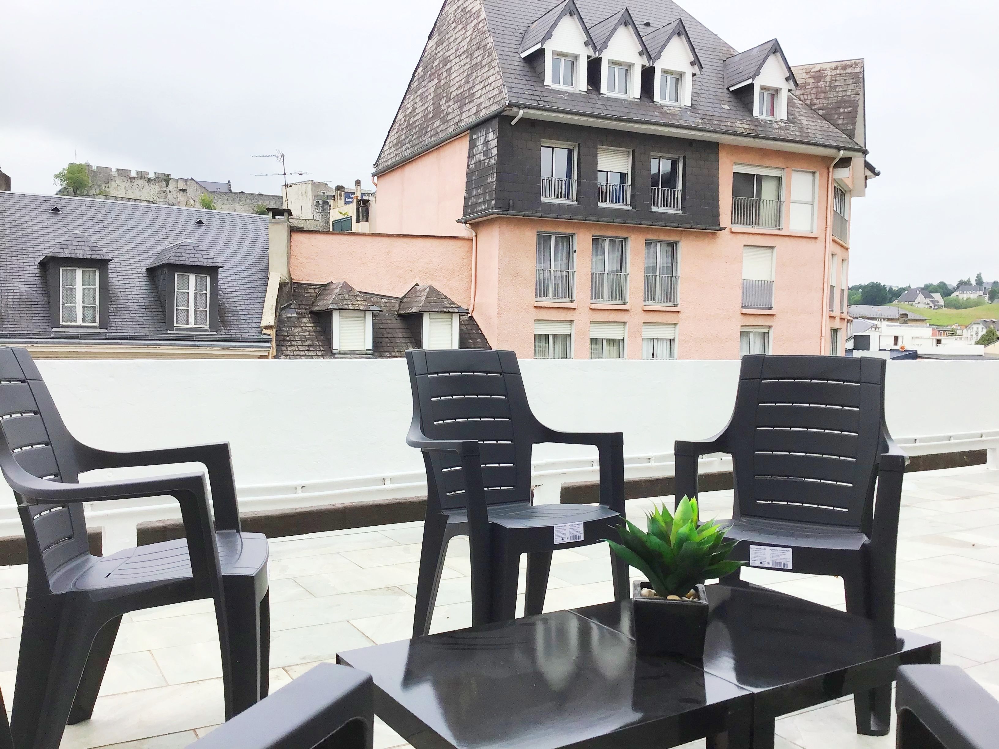Una terraza con sillas de plástico negro y una mesa, con vista a un edificio de fachada rosa y techo gris.