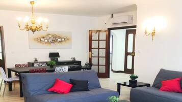 A modern living room with a grey sofa, a dining table, and a chandelier. There are two terraces, a private parking area, and four bedrooms.