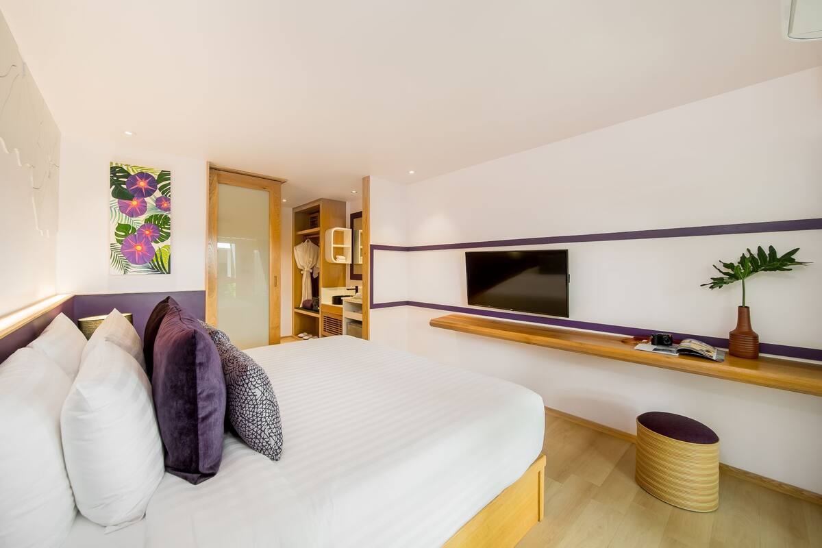 A modern hotel room with a bed, a TV, a plant, and a small stool.