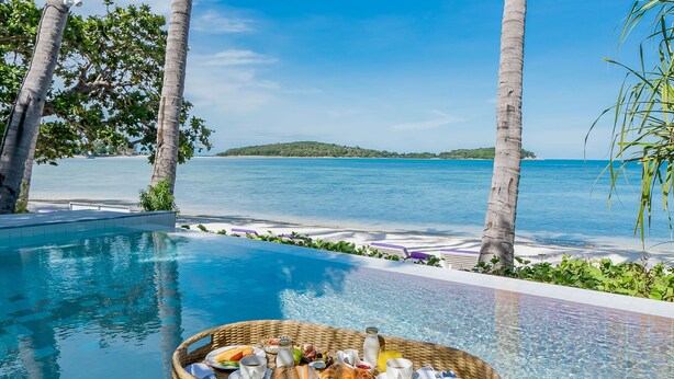 A poolside breakfast setup with a wicker basket filled with food and drinks, surrounded by palm trees and a clear blue sky. - La Vida Samui (Koh Samui)