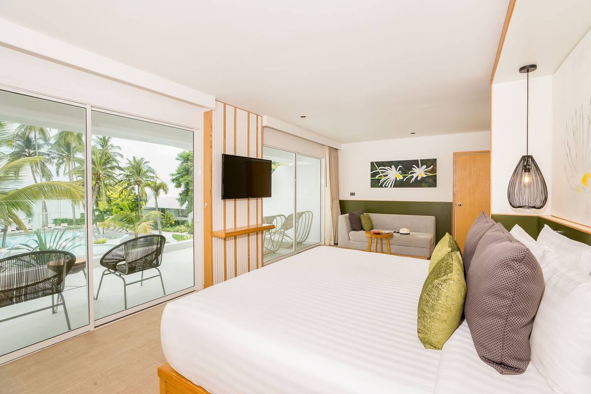 A modern hotel room with a large bed, a TV, a balcony with chairs, and a view of palm trees.