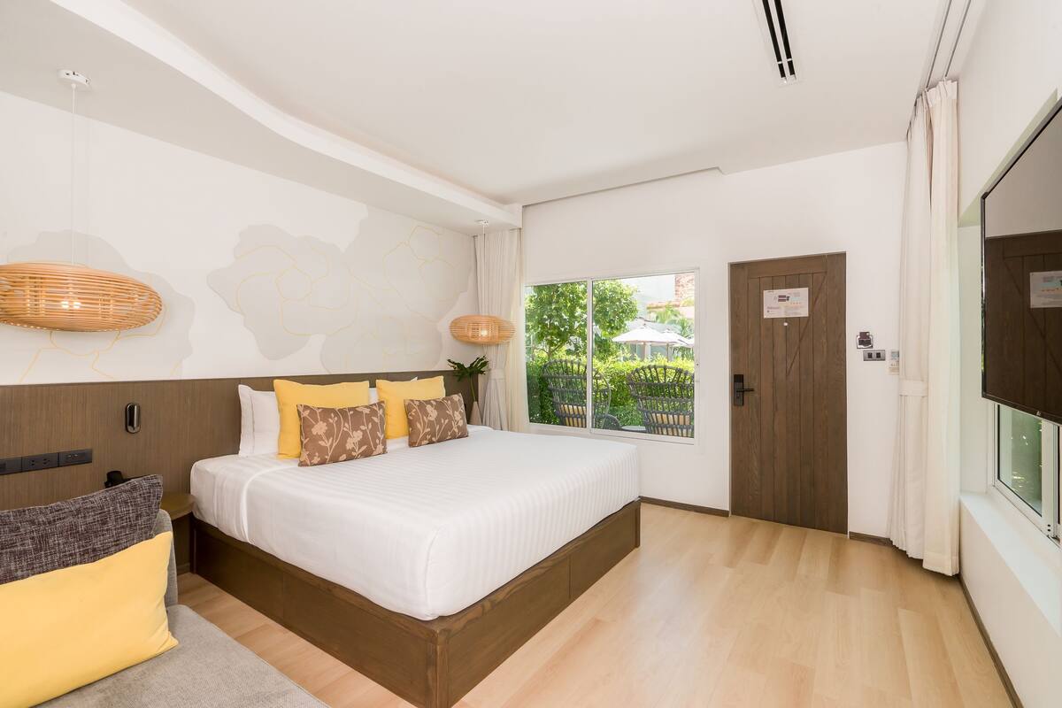 A modern bedroom with a large bed, a wooden headboard, a bedside table, and a window with a view of greenery.