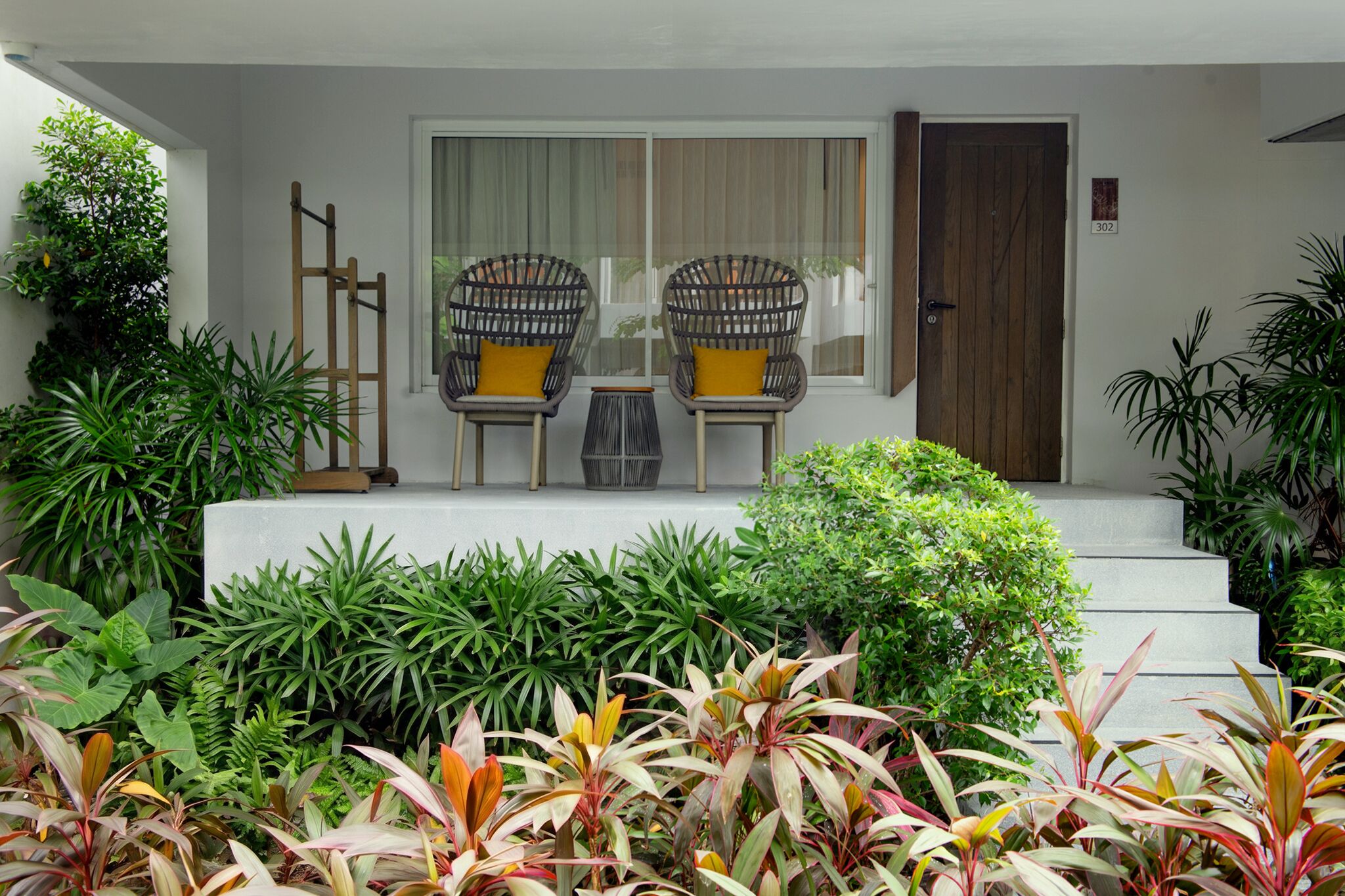 A house entrance with a wooden door, two wicker chairs with cushions, a small table, and a variety of plants.