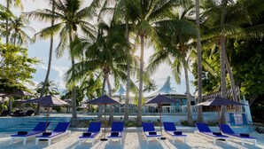 A beach with sun loungers and umbrellas, palm trees, and a building in the background. - La Vida Samui (Koh Samui)