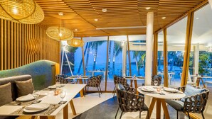 A modern restaurant with wooden ceiling panels, wicker pendant lights, and large windows offering a view of palm trees and the ocean. - La Vida Samui (Koh Samui)