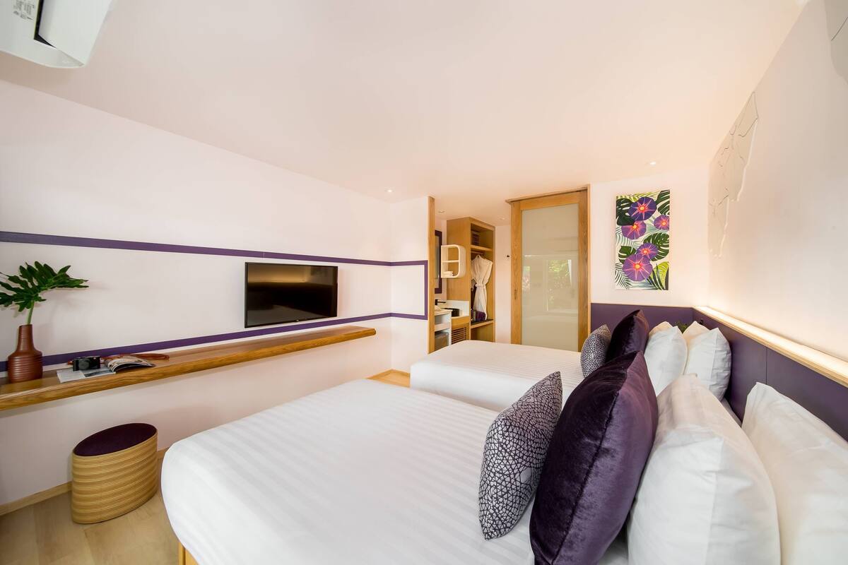 A modern hotel room with a large bed, a flat-screen TV mounted on the wall, a wooden bedside table with a plant, and a colorful artwork on the wall.