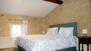 Un dormitorio con pared de piedra, una cama con ropa de cama floral, una ventana con cortinas y una mesita de noche con lámpara.