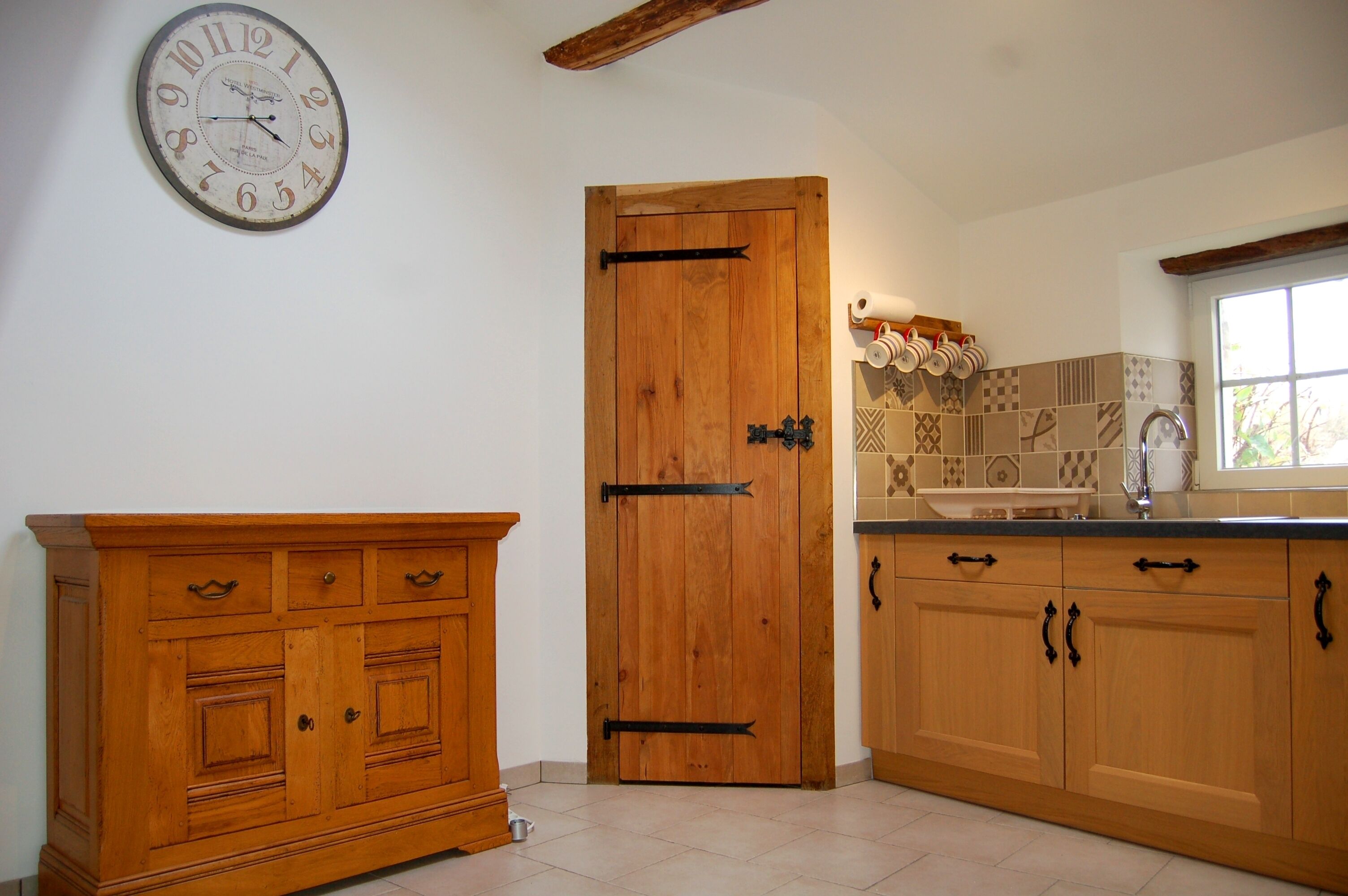 Une cuisine avec des armoires en bois, une porte en bois, une horloge murale et une fenêtre.